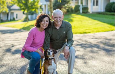 Senior white haired man posing with brunette woman outside Peconic Landing on a walk with their dog