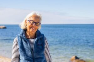 Senior woman with white hair and black glasses standing on the beach with the ocean behind her