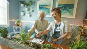 Short term respite care room, senior woman and nurse sitting on couch smiling doing an activity together